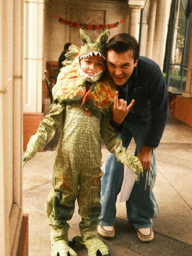 Javi crouched beside a young cast member in a dinosaur costume, both throwing a rock sign