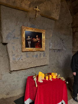 A cave wall set with a gilt-framed painting above an altar dressed in red cloth and candles